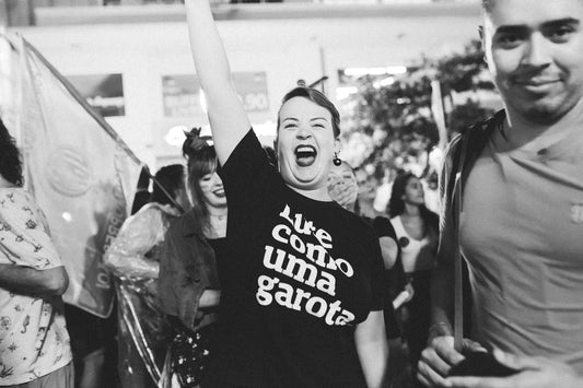foto preto e branca de uma mulher em uma manifestação com uma camiseta preta com a frase lute como uma garota em branco. A mulher é branca, de cabelos curtos e castanhos. Ela está gritando, com a mão direita erguida. Ao fundo, pessoas e bandeiras.