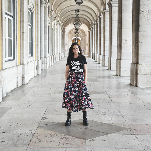 Foto colorida da Nathalia, mulher branca com cabelos castanhos, lisos e médios vestida com uma peita preta com a frase Lute como uma garota em branco e saia florida. Ela está no Arco do Triunfo da Rua Augusta em Lisboa.