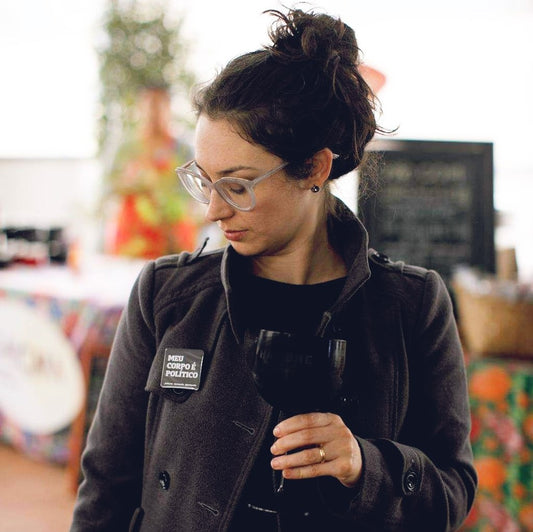 foto colorida da Lívia, mulher branca com cabelos castanhos, presos em coque. Ela está de blazer cinza escuro com botton quadrado preto com a frase Meu corpo é político em branco, olhando para ele e segurando uma taça preta.