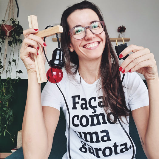 foto colorida em ambiente interno de uma mulher branca, com cabelos castanhos, lisos e compridos, usando óculos de armação marron e redonda, vestida com uma peita branca com a frase faça como uma garota em preto.