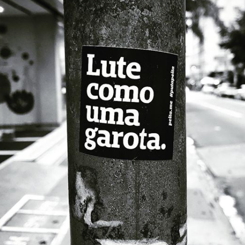Foto preta e branca de um adesivo preto com a frase lute como uma garota em branco, colado em um poste metálico.
