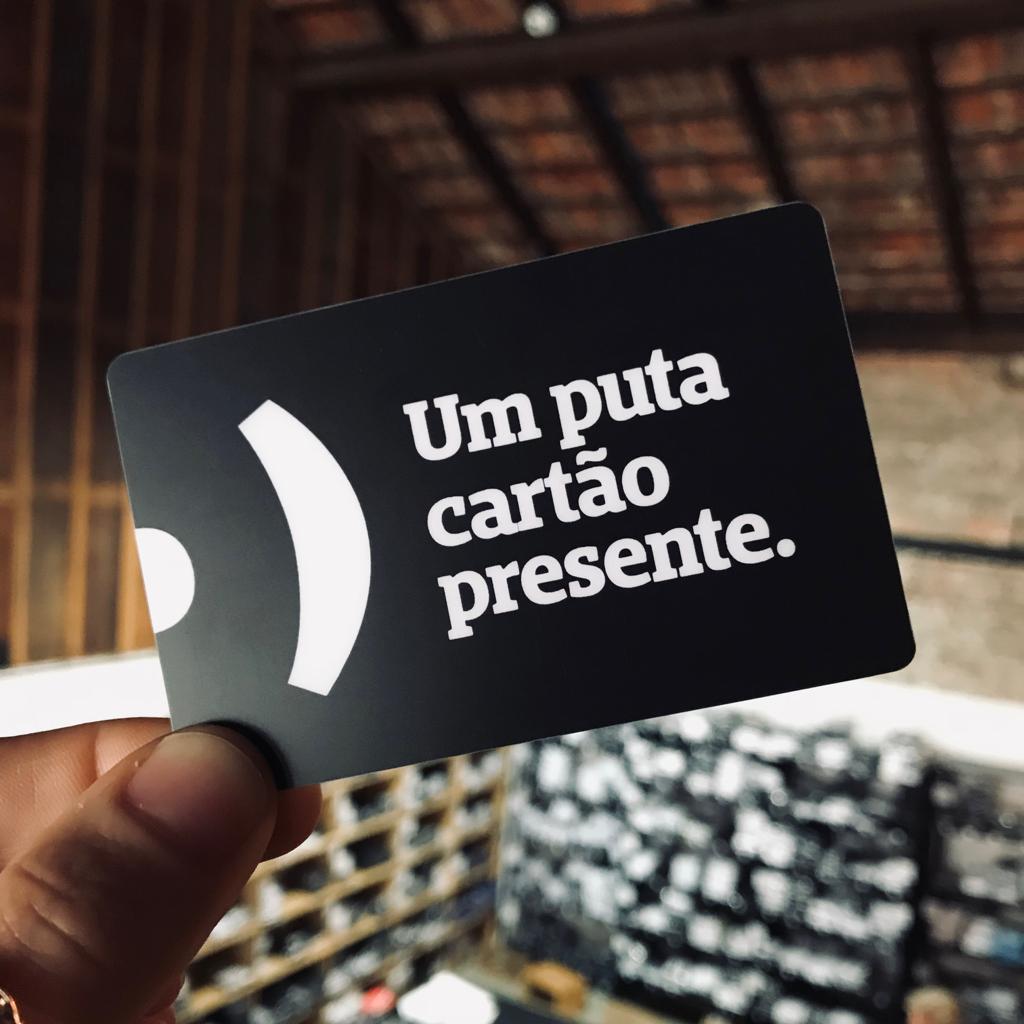 Foto colorida de um cartão preto com a logo da Peita, com a frase um puta cartão presente em branco. Está sendo segurado por uma mão branca.
