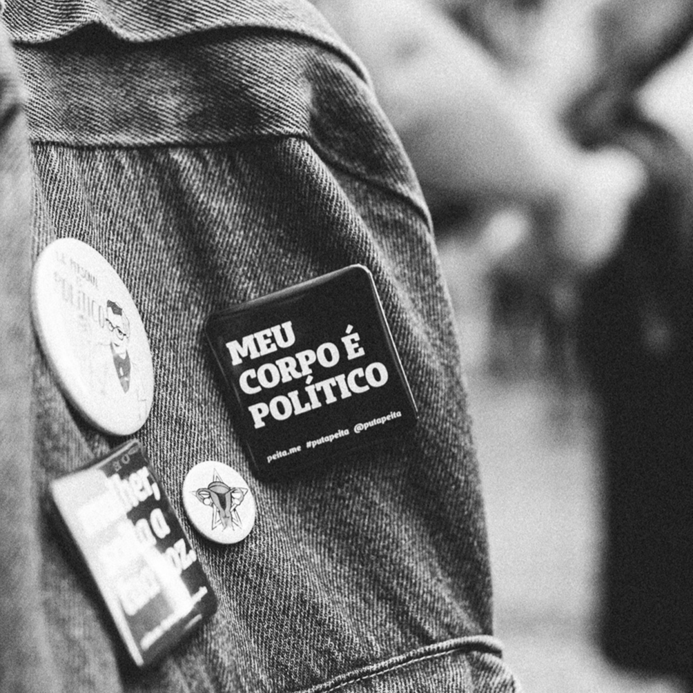 Foto preta e branca de uma mochila jeans com um botton preto, quadrado, com a frase meu corpo é político em branco.