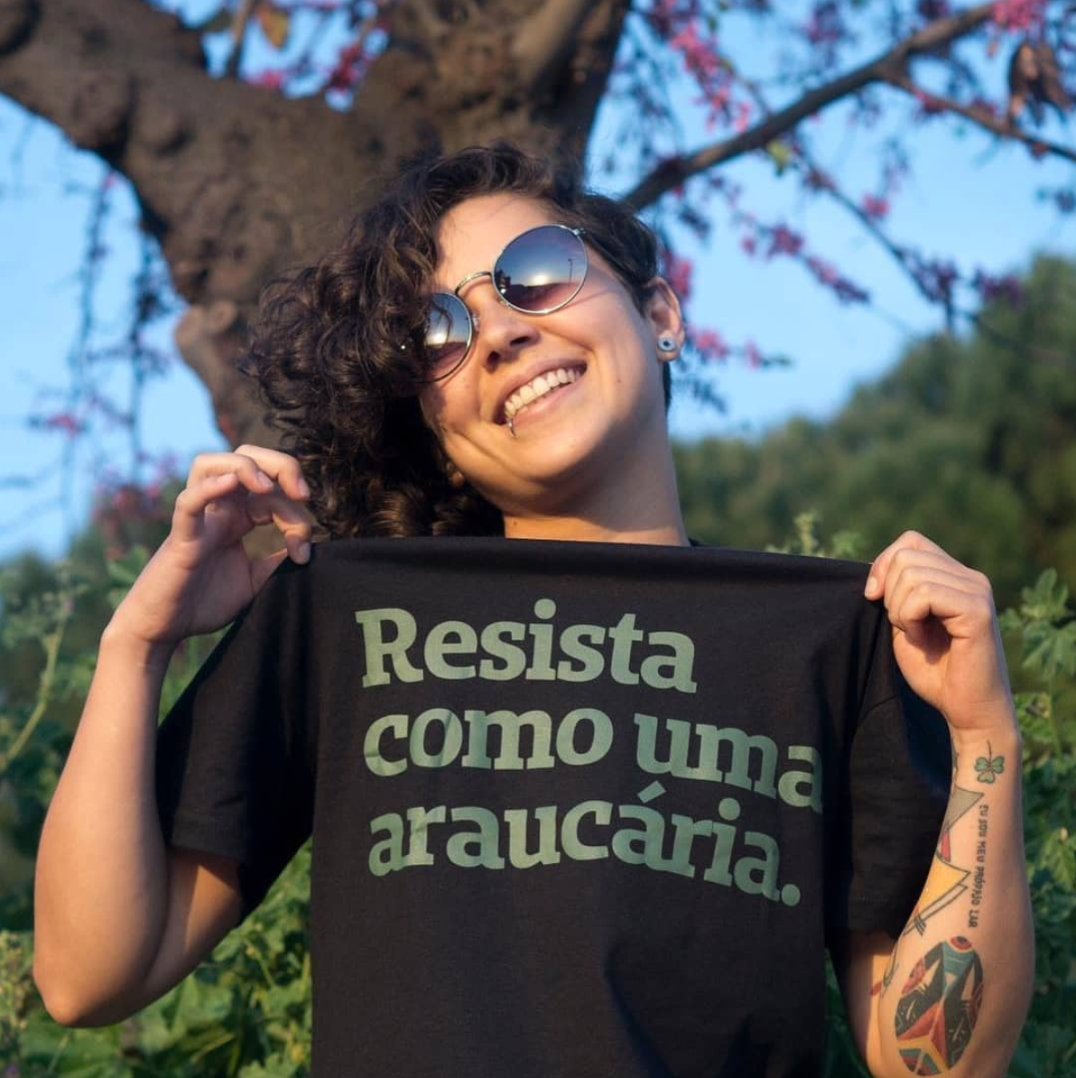 Foto colorida de uma mulher branca, cabelos castanhos, cacheados, curtos, com óculos escuros, piercing na boca, vestida com camiseta preta escrita resista como uma araucária em verde. Ela está puxando a estampa para cima, sorrindo.