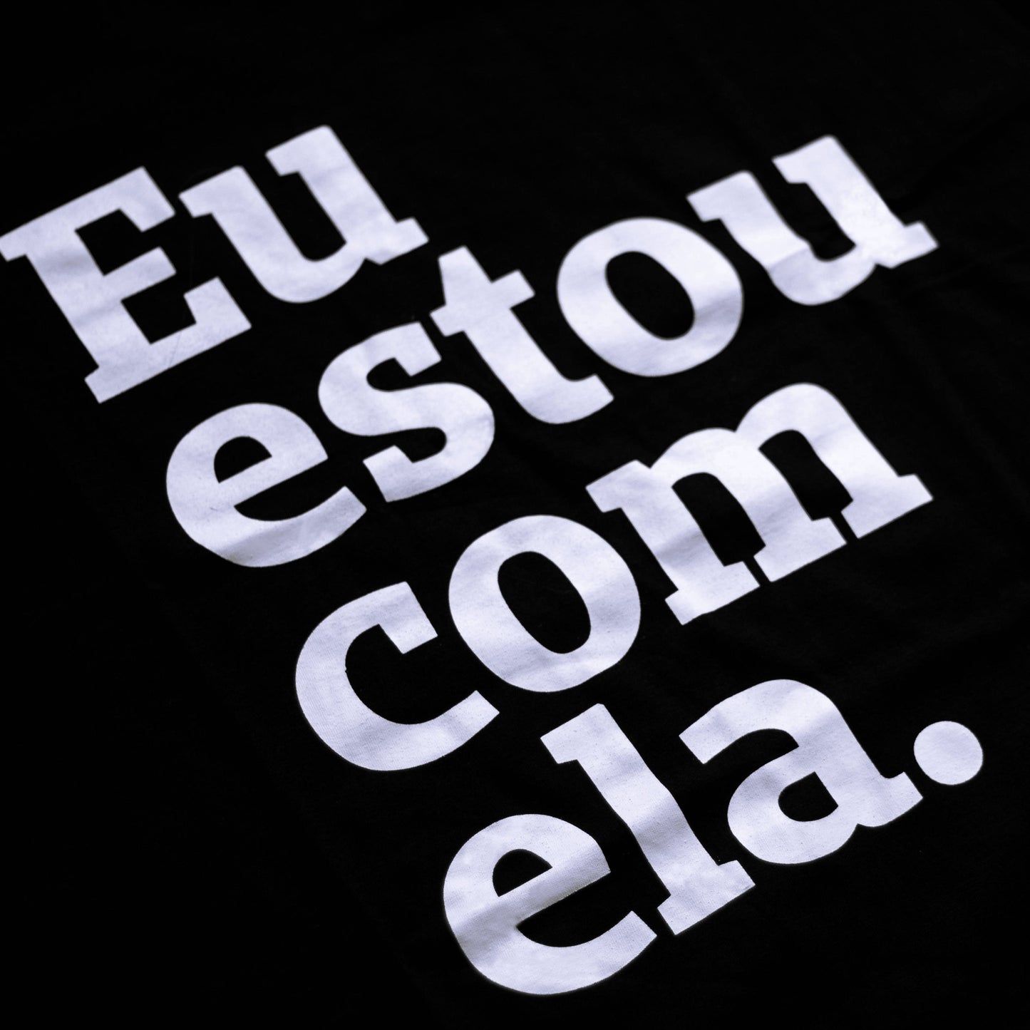 Foto preta e branca de uma camiseta preta com a frase eu estou com ela. Close na estampa.