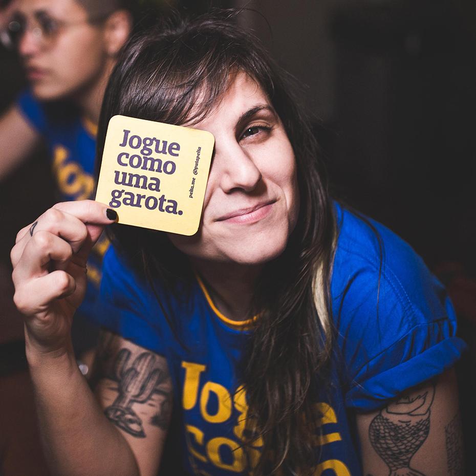Foto colorida de uma mulher branca de cabelo castanho, liso, com franja e veste uma camiseta azul escrito jogue como uma garota em amarelo. Ela segura um porta copo amarelo escrito jogue como uma garota em azul, posicionado em frente ao seu olho esquerdo. Tem tatuagens no braço e está sorrindo.