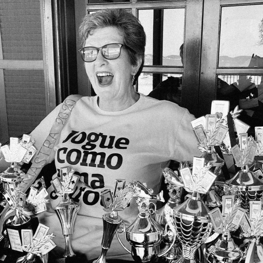 Foto preta e branca de uma mulher branca de cabelos curtos, lisos, claros e veste uma camiseta branca escrita jogue como uma garota em preto. Ela está sentada com vários troféus e medalhas a sua volta, sorrindo pra foto.