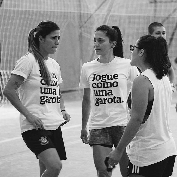 Foto preta e branca de 3 mulheres brancas. A do meio tem cabelos escuros, lisos, médios, presos em rabo. A da esquerda tem cabelos lisos, escuros nas raízes e loiros nas pontas. A da direita tem cabelo liso, escuro, preso em rabo, usa óculos. Ambas vestem peita branca com a frase jogue como uma garota em preto.