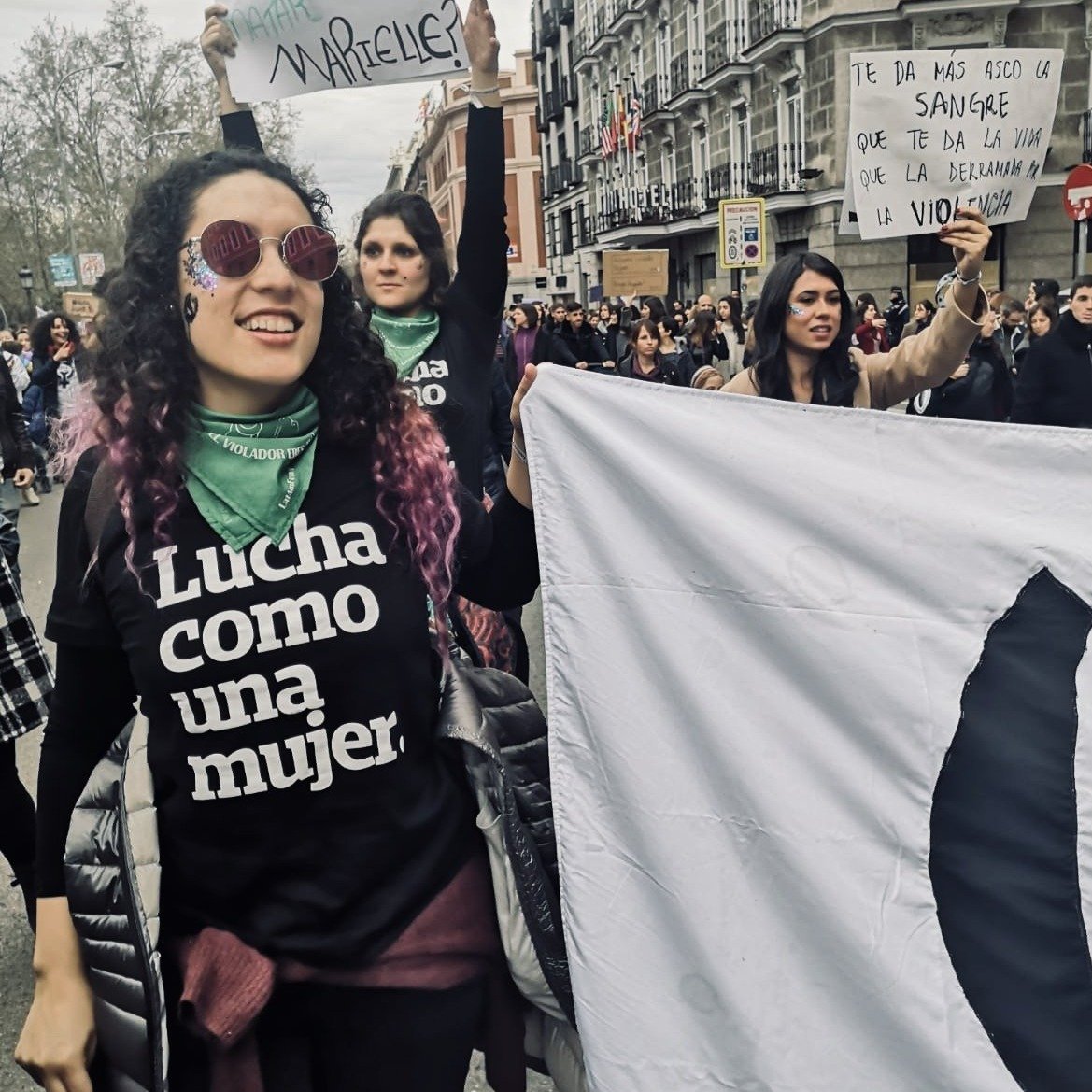 Foto colorida de mulher branca com cabelos castanhos, pontas rosas, cacheados, longos, com óculos rosa, vestindo camiseta preta escrita lucha como una mujer em branco. Ela está segurando uma bandeira com a logo da Peita, sorri para foto.