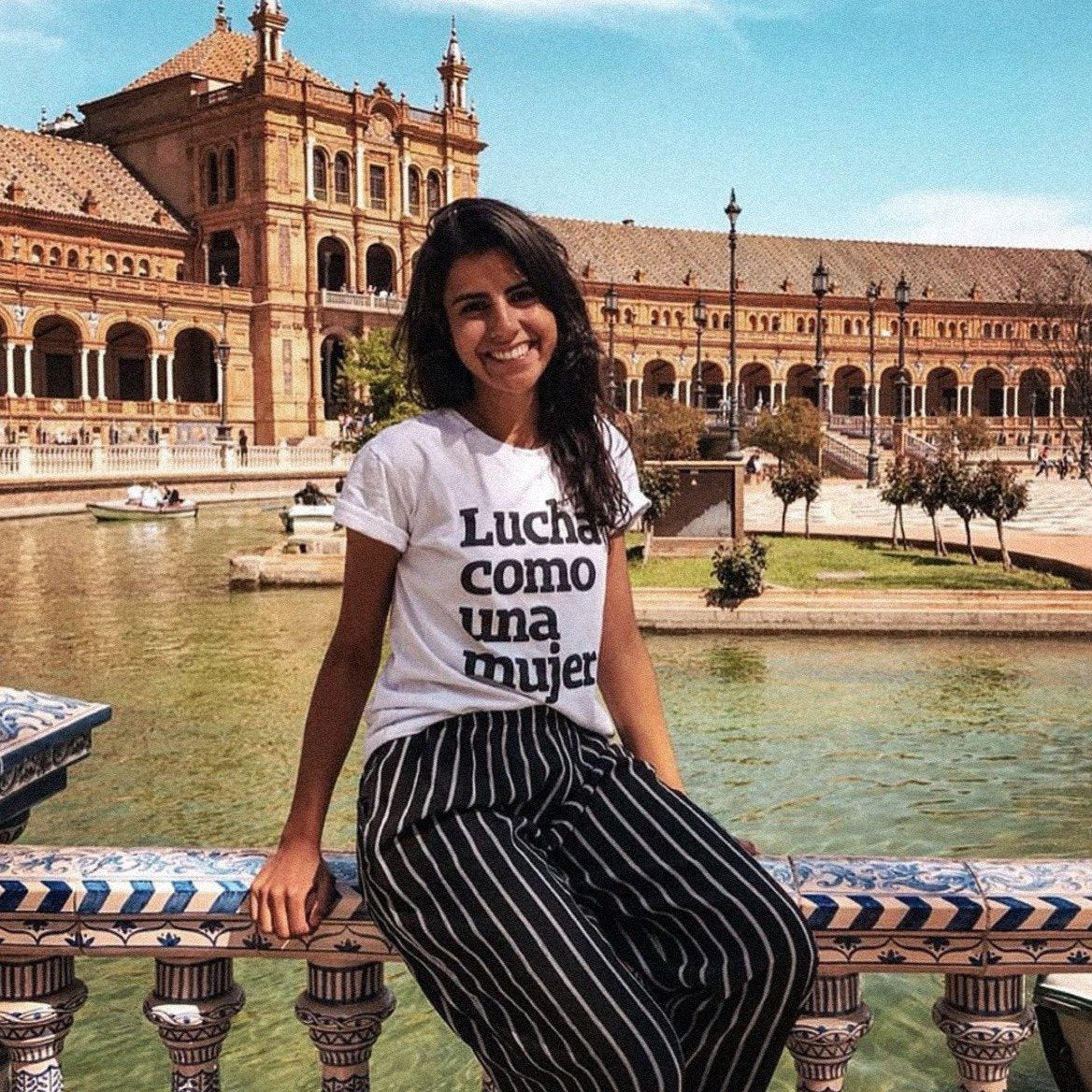 Foto colorida de uma mulher branca, com cabelos castanhos, lisos, médios, com camiseta branca escrita lucha como una mujer em preto. Está sentada em um corrimão, sorrindo para foto.
