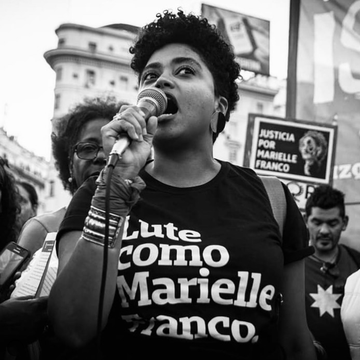 Foto branca e preta de uma mulher negra, cabelos crespos, pretos, curtos, está vestida com camiseta preta escrito lute como Marielle Franco branco. No pulso esquerdo tem uma pulseira e um lenço amarrado, na mão direita segura o microfone.