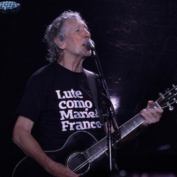 Foto colorida do Roger Waters, homem branco, com cabelos , grisalhos, com camiseta preta escrito lute como Marielle Franco em branco, toca um violão, enquanto canta no microfone.