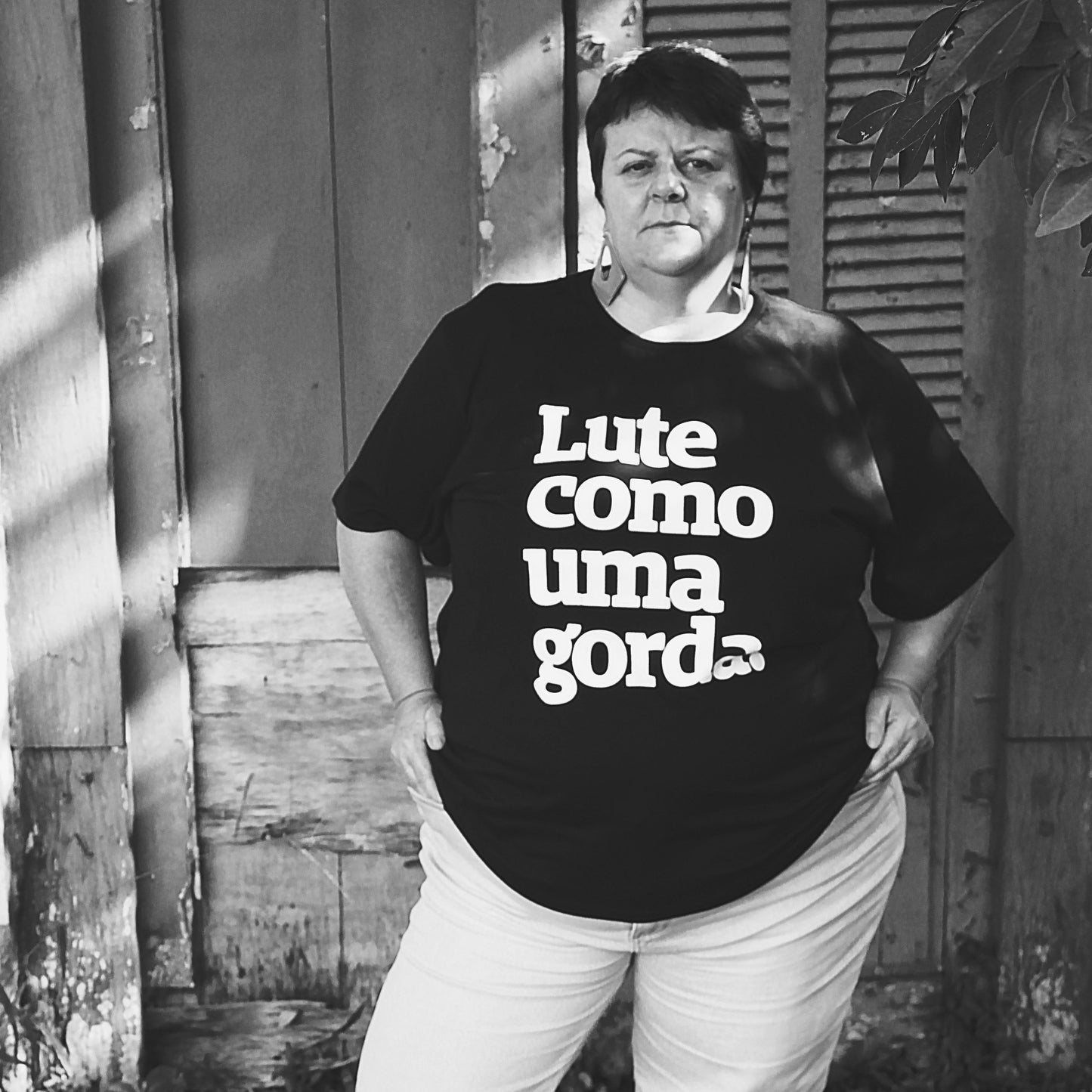 Foto preta e branca de uma mulher branca, cabelos lisos, soltos, curtos, castanhos. Está vestida com uma camiseta preta escrito lute como uma gorda embranco . Está séria, com as mãos na cintura.