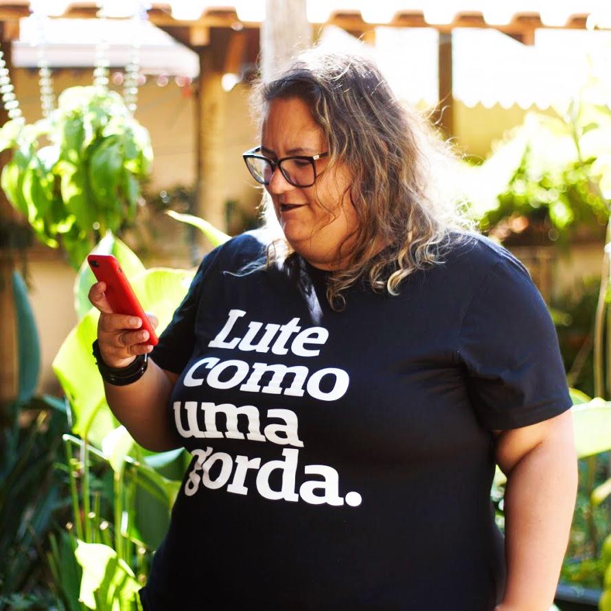 Foto colorida de uma branca, cabelos lisos, soltos, castanhos claros, na altura dos ombros. Está uma mulher vestida com camiseta preta escrito lute como uma gorda em branco. olha para o celular em sua mão direita.