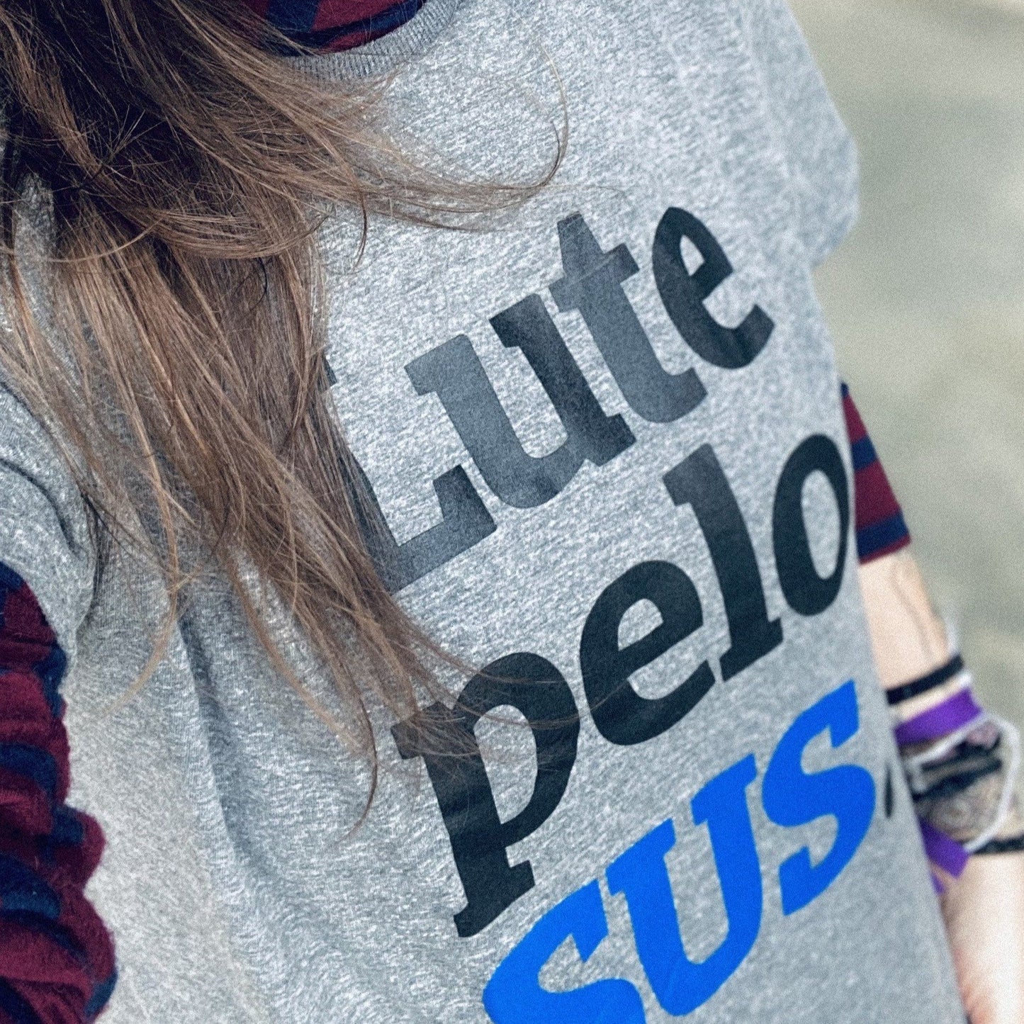 Foto selfie colorida do tronco de uma mulher branca, de cabelos castanhos, longos, lisos, está vestida com camiseta cinza escrita lute pelo sus em preto e azul. usa uma blusa de manga longa, com listras vermelhas e azuis por baixo.