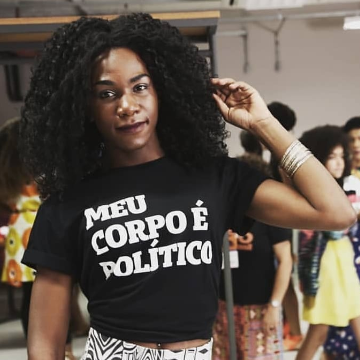 Foto colorida de uma mulher negra, cabelos cacheados, pretos, médios, soltos, usa pulseira no pulso esquerdo, está vestida com camiseta preta escrita meu corpo é político em branco. Segura os cabelos com a ponta dos dedos.