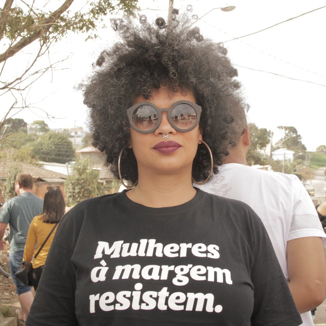 Foto colorida de uma mulher negra, cabelos castanhos, cacheados, curtos, soltos, usa brincos de argolas prateadas, óculos escuros com armação preta, piercing no nariz, está vestida com camiseta preta escrita mulheres à margem resistem em branco.