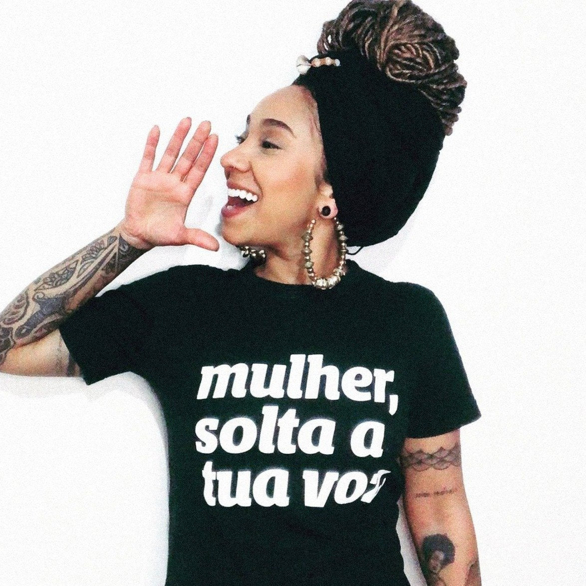Foto colorida de uma mulher negra, cabelos com dreads castanhos claros, presos com turbante preto, usa brincos de argola, tem tatuagens nos braços, está vestida com camiseta preta escrita mulher, solta tua voz em branco. Ela está olhando para esquerda com a mão aberta fingindo gritar.