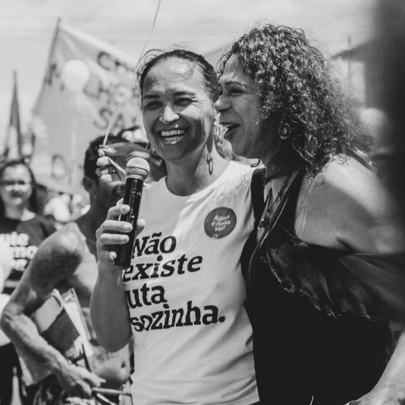 Foto colorida de 2 mulheres: a da direita, mulher branca, cabelos castanhos, lisos, presos em rabo, está vestida com camiseta branca escrita não existe luta sozinha em preto. Ela segura um microfone e sorri, abraça dá a mulher negra, cabelos cacheados, castanhos, médios com blusa preta.