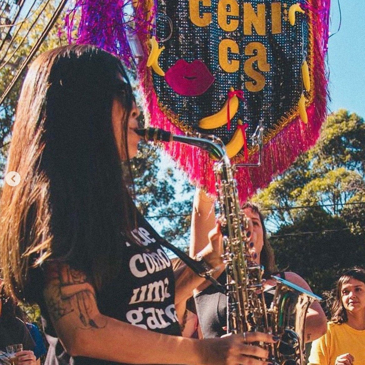 Foto colorida de uma mulher branca, cabelos castanhos, lisos, longos, com tatuagem no braço direito, óculos escuros, usa camiseta preta escrita toque como uma garota em branco. Ela está tocando um saxofone em um bloco.