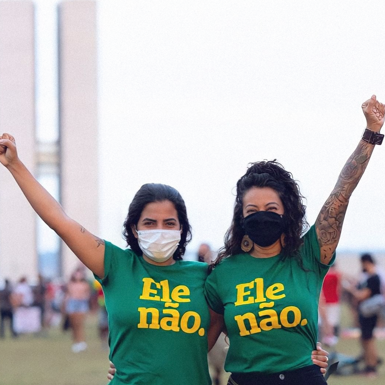 Foto colorida em ambiente externo de duas mulheres em frente ao Congresso Nacional com o punho erguido vestidas com uma peita verde escrito Ele não em amarelo. Ambas são brancas, tem cabelos castanho e presos. A da esquerda usa máscara branca e a da direita usa máscara preta e tem braço erguido tatuado. Ao fundo uma manifestação contra o presidente.