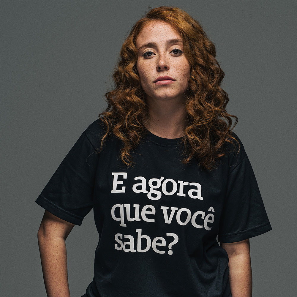 Foto colorida de uma mulher branca, com os cabelos cacheados soltos, médios e ruivo. Ela está vestindo uma camiseta preta escrito e agora que você sabe? em branco. Está olhando séria, com a mão esquerda no quadril.