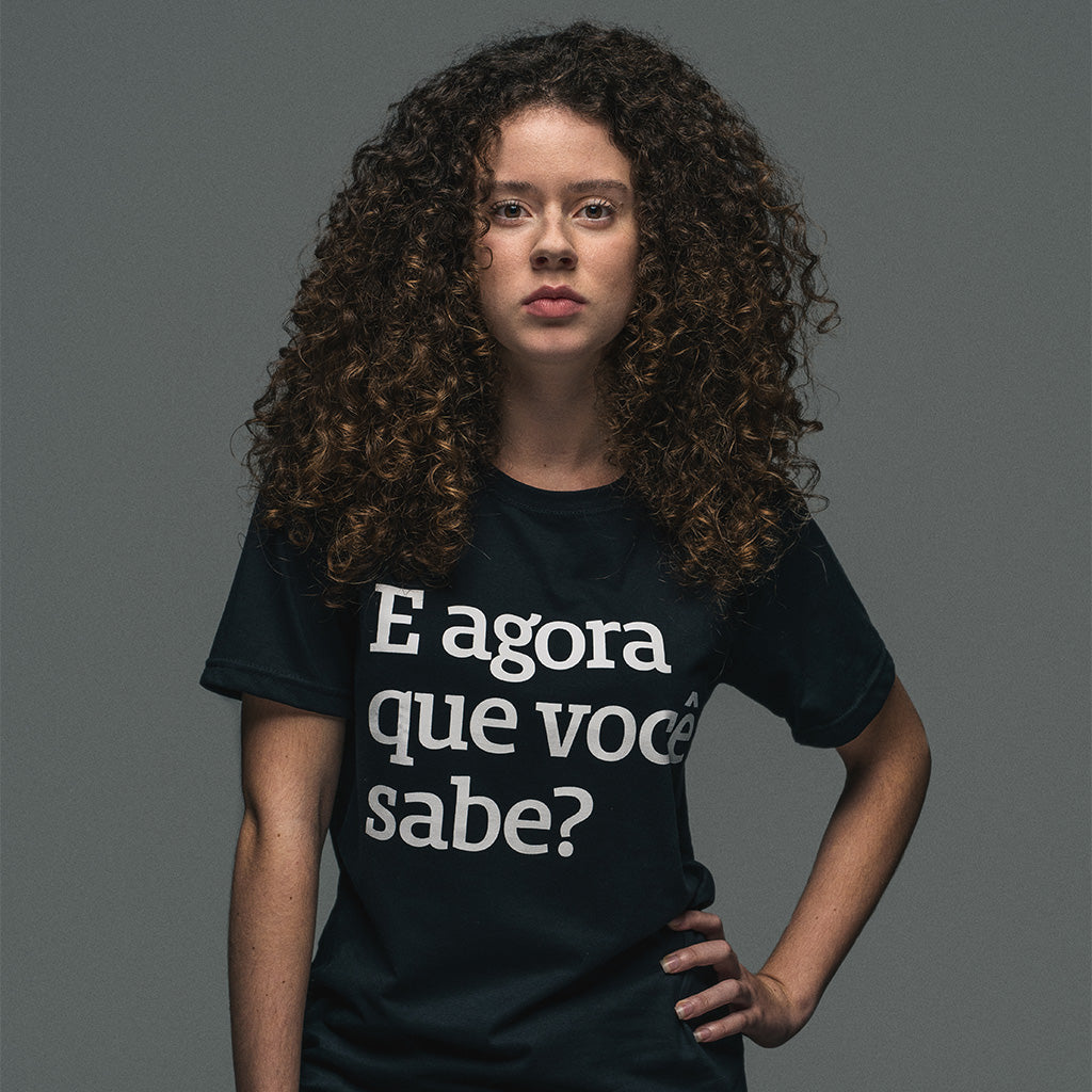 Foto colorida de uma mulher branca, com os cabelos cacheados soltos, médios e castanhos. Ela está vestindo uma camiseta preta escrito e agora que você sabe? em branco. Está olhando séria, com a mão direita no quadril.
