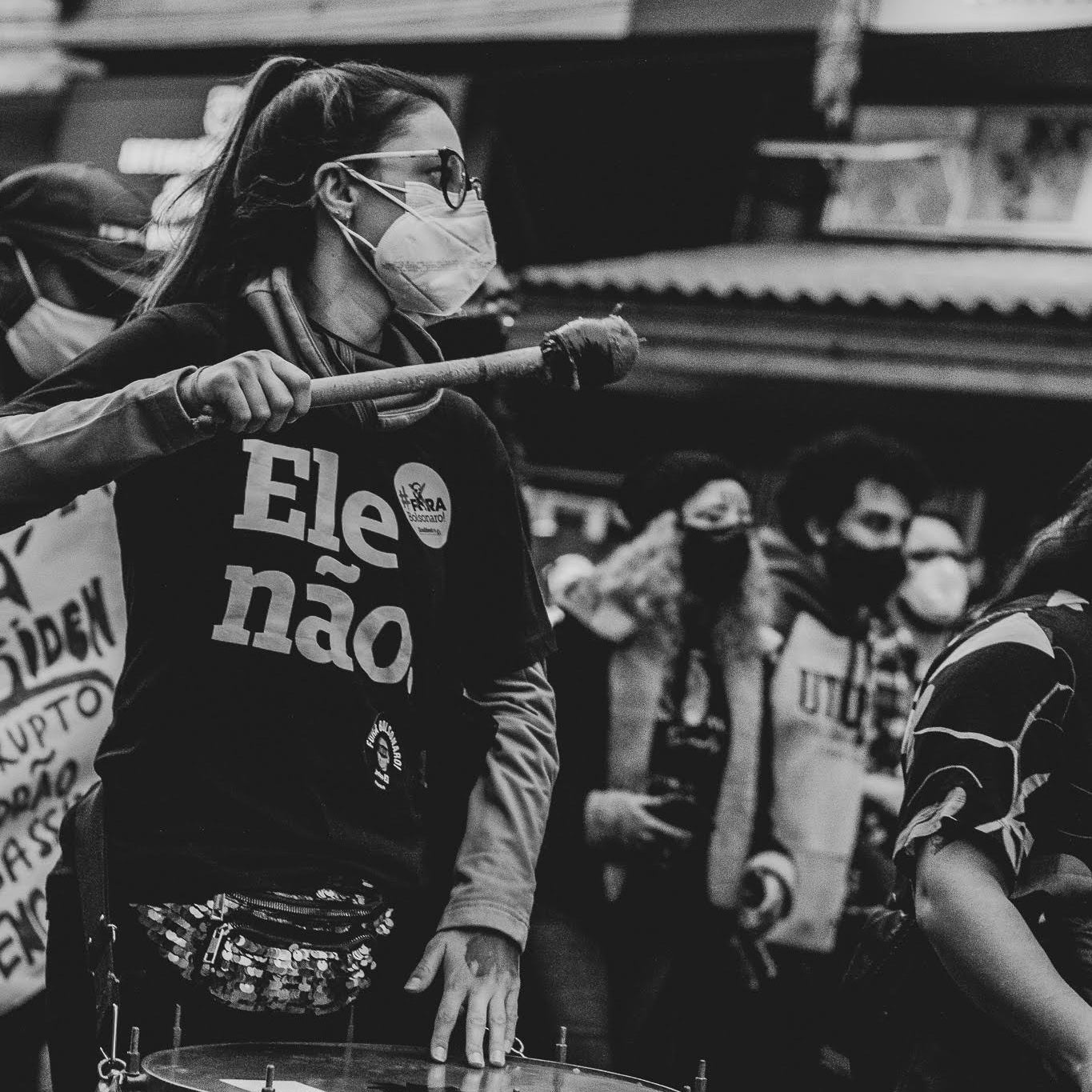Foto preto e branco de mulher branca com cabelos lisos, escuros, preso em rabo. Veste camiseta preta escrito ele não em branco, usa máscara branca e óculos. Toca um tambor. Fundo uma manifestação .