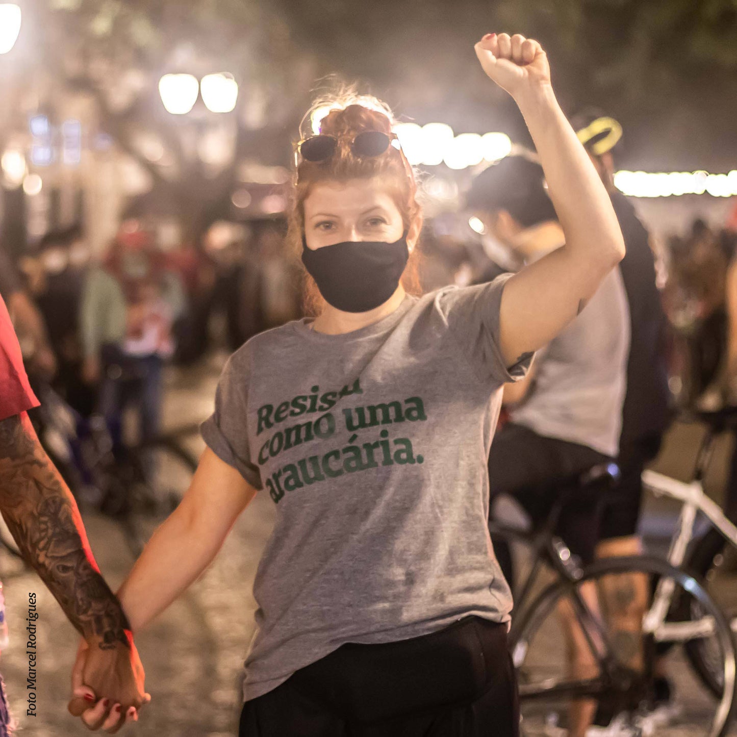 Foto colorida de uma mulher branca de cabelo ruivos lisos preso em coque, e óculos apoiado no topo da cabeça, veste camiseta cinza com frase resista como uma araucária em verde, com punho direito erguido e de mãos dadas com outra pessoa de braço tatuado. Fundo desfocado