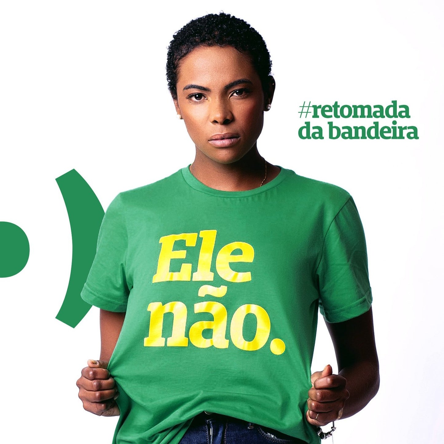 Foto colorida de uma mulher negra, de olhos escuros, cabelo raspado, veste camiseta verde escrito Ele não em amarelo. Ela esta séria, segurando a camiseta. Fundo branco