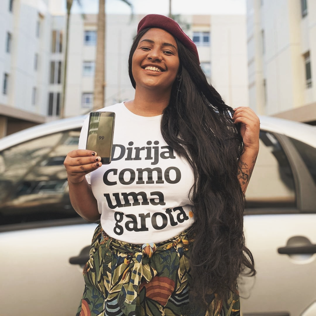 Foto colorida de uma mulher negra, cabelos lisos, soltos, longos e castanhos. Vestida com uma camiseta branca escrito dirija como uma garota em preto. Ela está sorrindo, mostrando o celular na mão direita, e a mão esquerda está no cabelo.