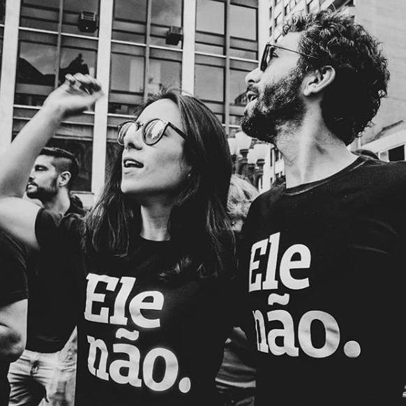Foto preto e branca de uma mulher branca, cabelos lisos, castanhos e compridos e um homem branco, com cabelos curtos, castanhos e barba. Ambos estão usando uma peita preta com a frase ele não em branco. Ela está com o punho do braço direito levantado.