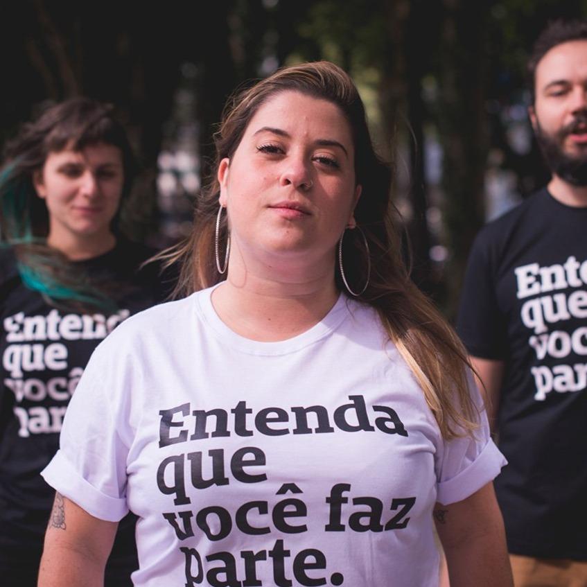 Foto colorida de 2 mulheres brancas, com cabelos lisos, compridos, castanho e loiro e 1 homem branco com cabelos lisos, curtos, castanhos e barba curta . A do meio está de camiseta branca escrito entenda que você faz parte em preto. , A mulher da sua esquerda e o homem,vestem camisetas pretas com a frase entenda que você faz parte em branco Todas olham sério pra foto.