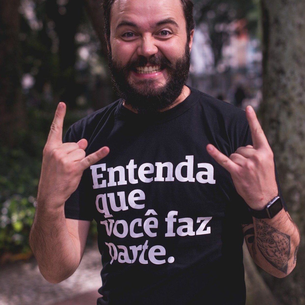 foto colorida de um homem branco, cam cabelos e barba castanhos. Está com camiseta preta escrita entenda que você faz parte em branco. Ele está fazendo uma careta, sorrindo, fazendo um sinal de rock'n roll com a mão.
