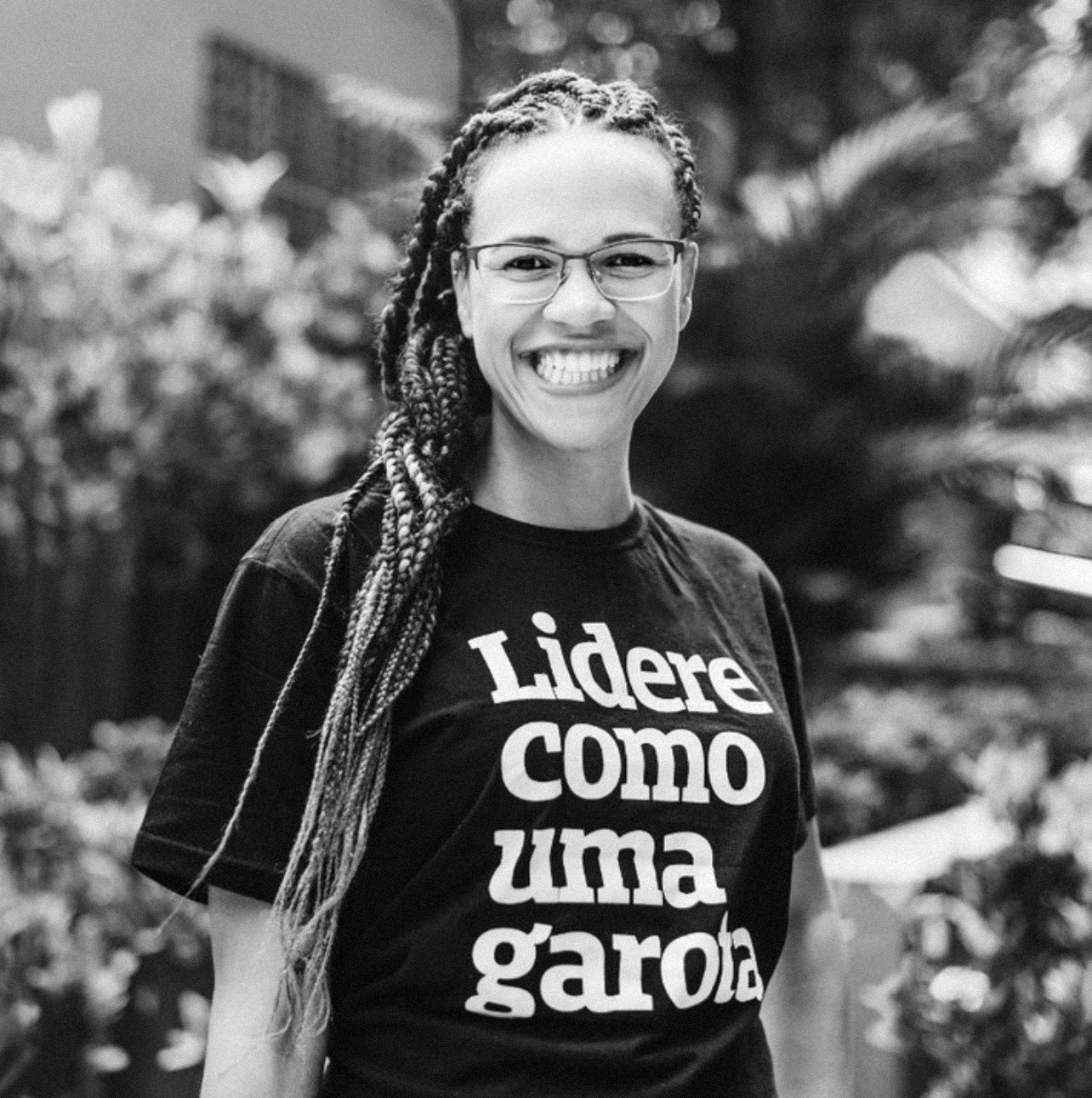Foto preto e branco de uma mulher negra com cabelos trançados e longos, veste uma camiseta preta escrito Lidere com o uma garota em branco. Usa óculos de grau e sorri. Fundo desfocado