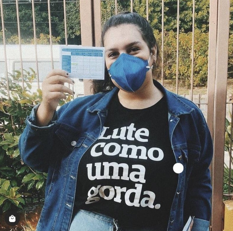 Foto colorida de mulher branca de cabelos escuros preso em rabo. Veste camiseta preta com frase Lute como uma gorda em branco e jaueta jeans por cima. Usa mascara azul e segura carteira de vacinação com a mão esquerda. Fundo portão e plantas
