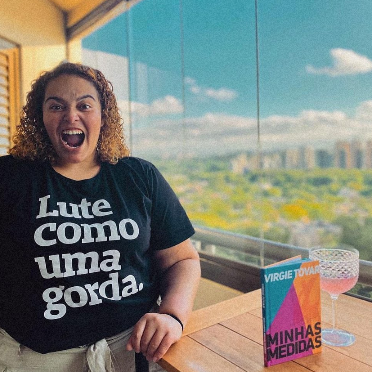 Foto colorida de mulher branca de cabelos claros, cacheados, curtos e soltos. Veste uma camiseta preta com frase Lute como uma gorda em branco. Apoia a mão em uma mesa de madeira que possui um livro de titulo Minhas medidas e uma taça com liquido rosa claro. Fundo janela de prédio alto desfocado