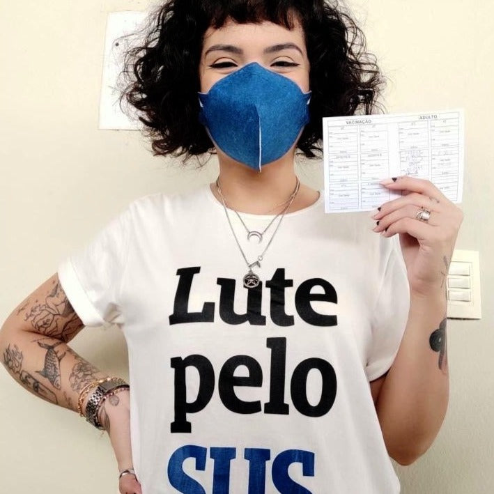 Foto colorida de uma mulher branca com cabelos castanhos, com corte chanel, veste uma camiseta branca escrito lute pelo sus em preto e azul e mascara azul. Segura com a mão direita a carteira de vacinação. Fundo branco