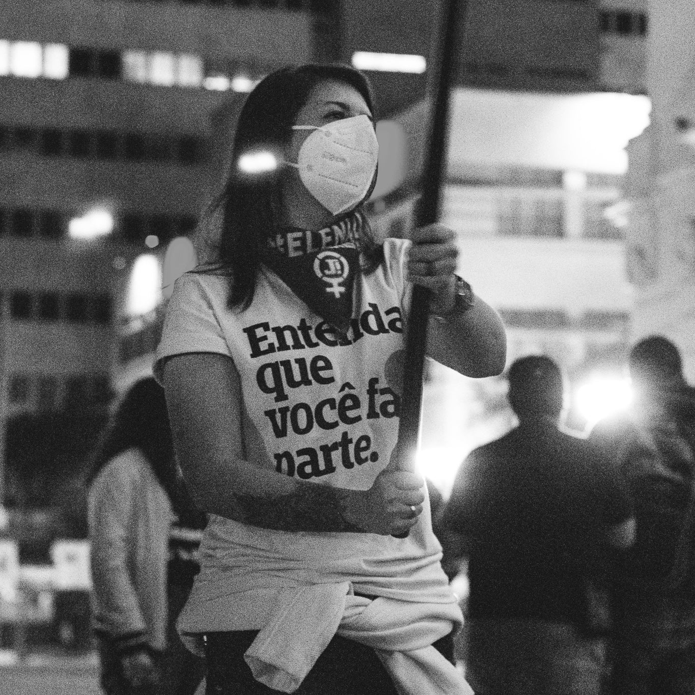 Foto preto e branco de uma mulher branca com cabelos escuros e curtos, veste uma camiseta branca escrito Entenda que você faz parte em preto. Usa máscara branca, lenço escrito ele não em branco e símbolo feminista, Segura com as duas mãos uma haste de bandeira e uma blusa branca amarrada na cintura. Fundo prédios e pessoas