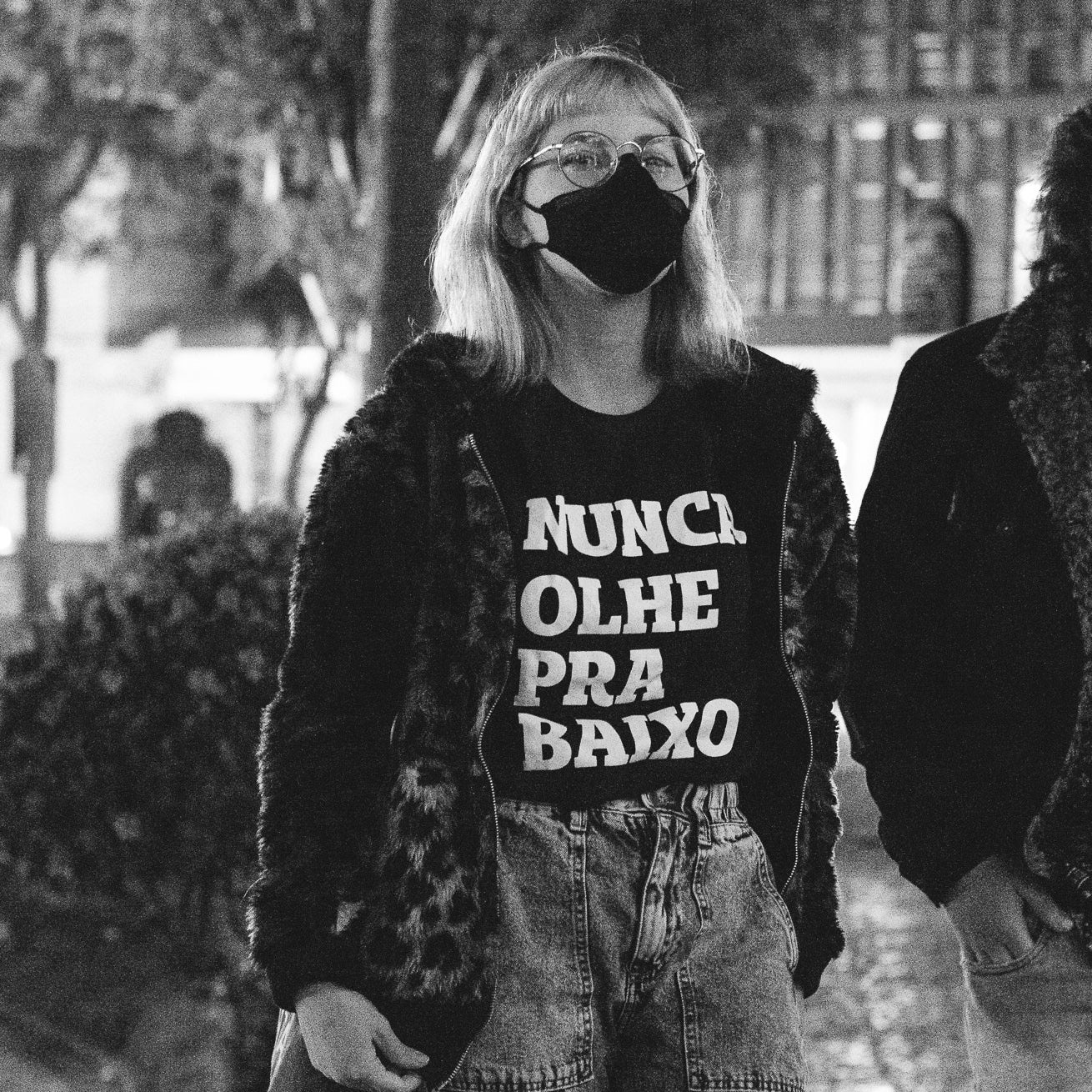 Foto preto e branco de mulher branca de cabelos claros curtos e soltos, veste camiseta preta com frase Nunca olhe pra baixo em branco, e uma jaqueta de pelos e estampa de onça, com mascara preta e óculos de grau redondos. Fundo desfocado