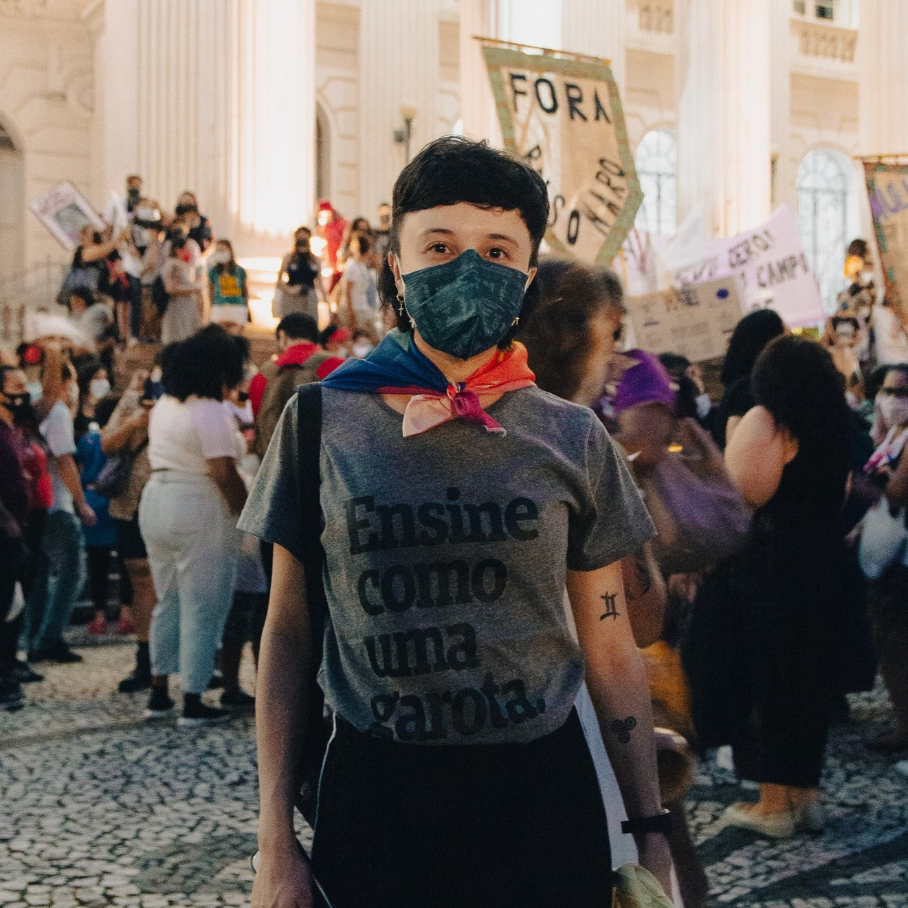 Foto colorida de uma mulher branca de cabelos escuros e curtos, veste uma camiseta cinza escrito Ensine como uma garota em preto. Usa um lenço colorido amarrado no pescoço e mascara verde escuro, com tatuagens no braço direito. Fundo manifestação.