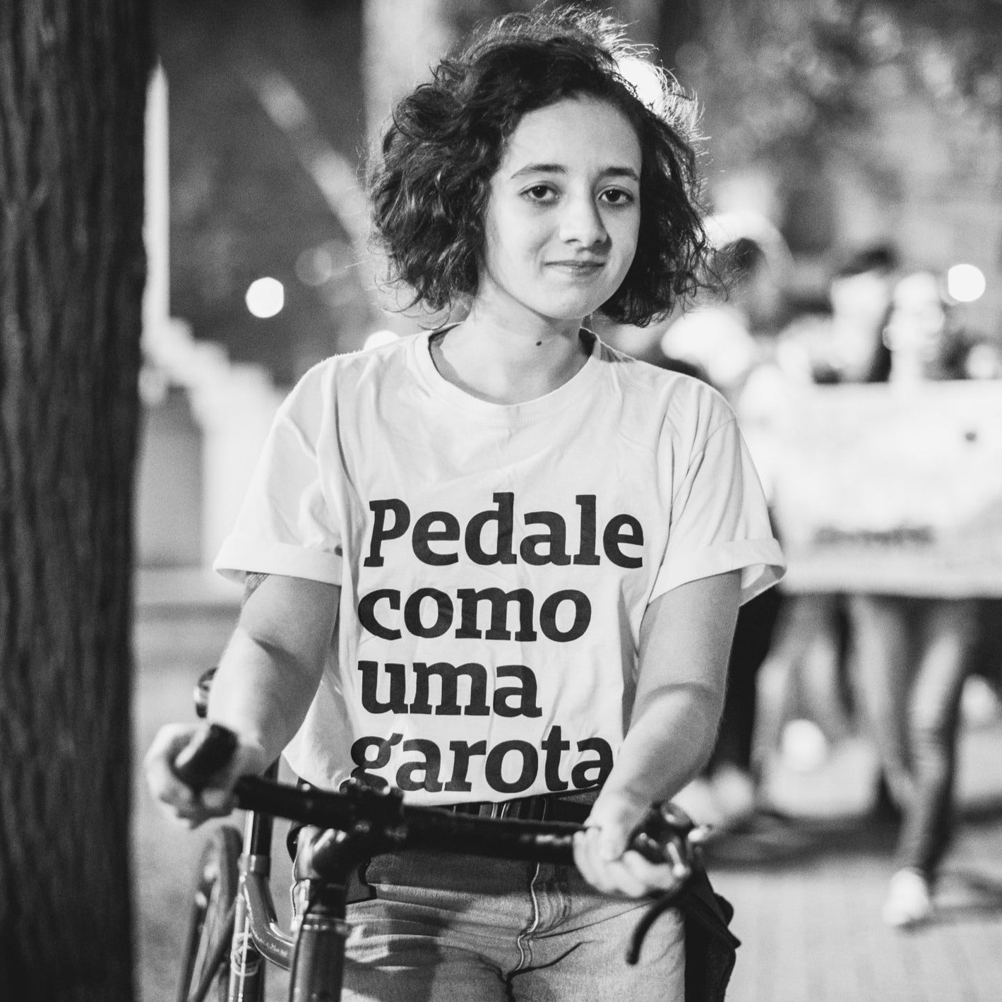 Foto preto e branco de uma mulher branca de cabelos castanhos encaracolados e curtos, veste uma camiseta branca com a frase Pedale como uma garota em preto, segura uma bicicleta ao lado do corpo. Fundo desfocado.