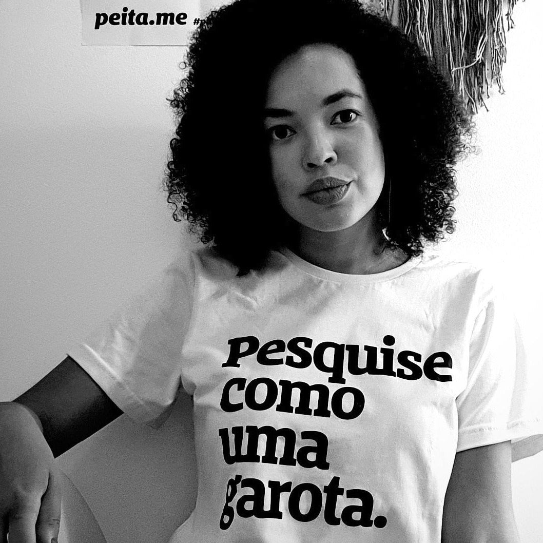 Foto preta e branca de uma mulher negra, cabelos crespos, pretos, soltos, chanel, vestida com camiseta branca escrita pesquise como uma garota em preto. Ela olha séria para foto.