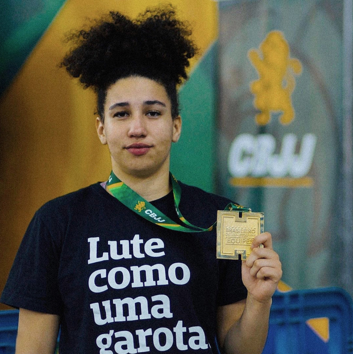 Foto colorida de Maria, mulher negra de cabelos crespo presos, medalhista em jiu jitsu, veste camiseta preta com frase Lute como uma garota em branco, segura com a mão direita uma medalha com cordão no pescoço. Fundo desfocado