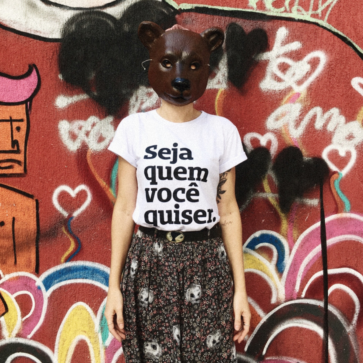 Foto colorida de mulher branca que veste uma camiseta branco com a frase Seja quem você quiser em preto, tem tatuagem no braço direito e usa calça estampada com flores. Usa uma máscara de urso marrom escuro que cobre o rosto todo. No fundo muro vermelho com desenhos coloridos.