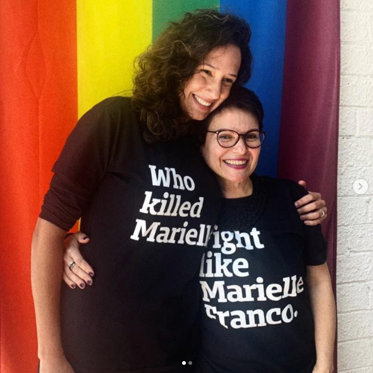 Foto colorida de 2 mulheres brancas, da esquerda é com cabelos castanhos, cacheados, médio, soltos, a da direita tem cabelos castanhos, lisos, curtos, soltos, usa óculos de grau. A da esquerda usa camiseta preta escrita who killed Marielle em branco e a da direita está com camiseta preta escrito fight like Marielle Franco em branco, Ambas estão sorrindo e tem uma bandeira LGBTI+ na parede.