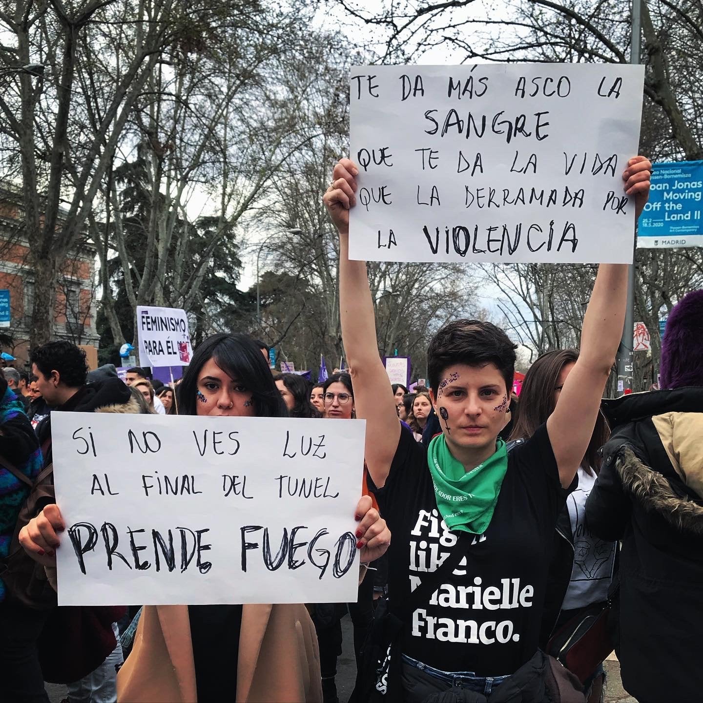 Foto colorida de 2 mulheres brancas, da esquerda tem cabelos pretos, lisos, médio, rosto pintado com 2 linhas verdes, casaco bege,e a da esquerda tem cabelos castanhos, lisos, curtos, com rosto pintado com símbolos do feminismo. A da direita está com camiseta preta escrito fight like marielle franco em branco, está segurando um cartaz pra cima, com bandana verde no pescoço. A Outra mulher segura um cartaz na altura do peito, está com casaco bege. Ambas olham sério para a foto no meio de um protesto.