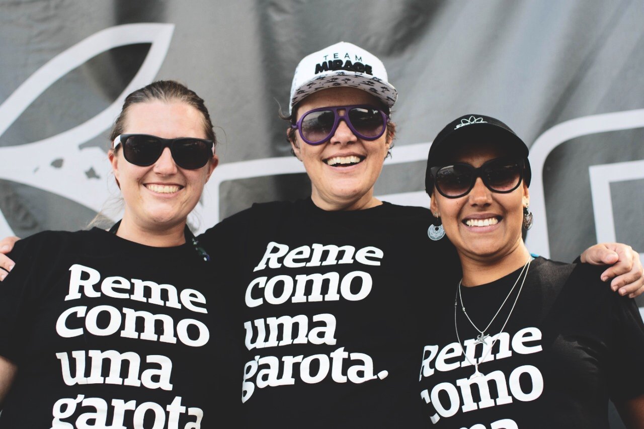 Foto colorida de 3 mulheres brancas, cabelos castanhos, presos, sendo que a do meio usa boné branco, estão vestidas com camiseta preta escrita reme como uma garota em branco, usam óculos escuros e sorriem para foto.