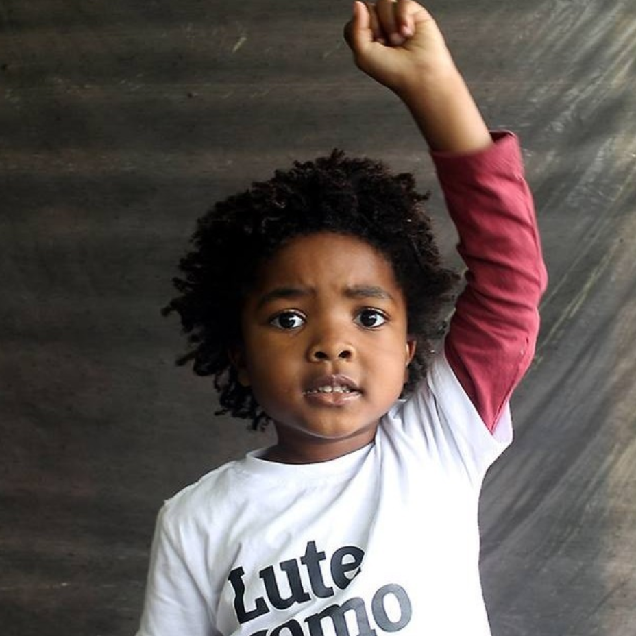 Foto colorida de uma menina negra, cabelos castanhos, crespos, curtos, vestida com camiseta branca escrita lute como uma garota em preto. Ela está com punho direito fechado levantado, olha séria para foto.