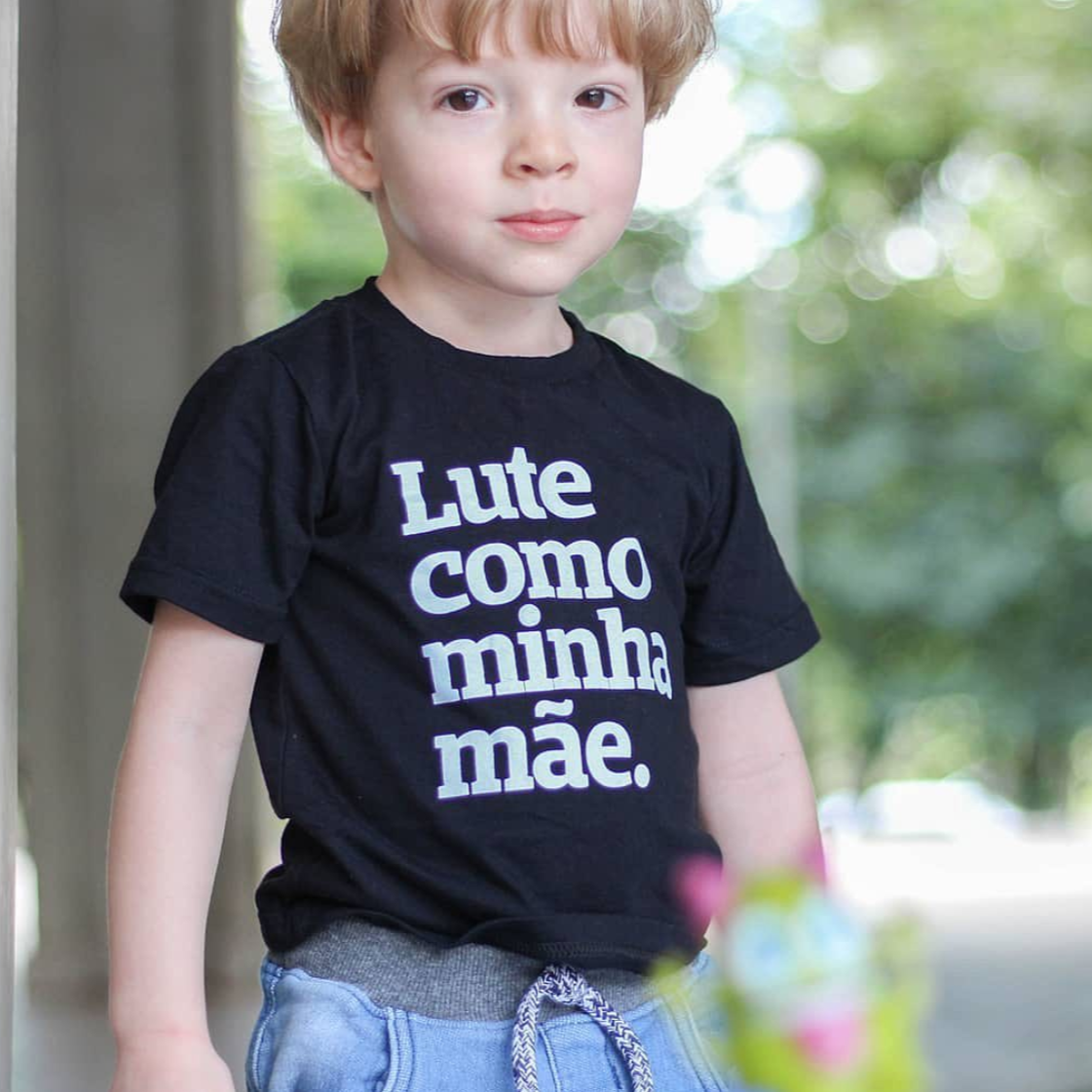 Foto colorida de um menino branco, cabelos loiros, curtos, vestido com camiseta preta escrito lute como minha mãe em branco. Olha sério para foto.